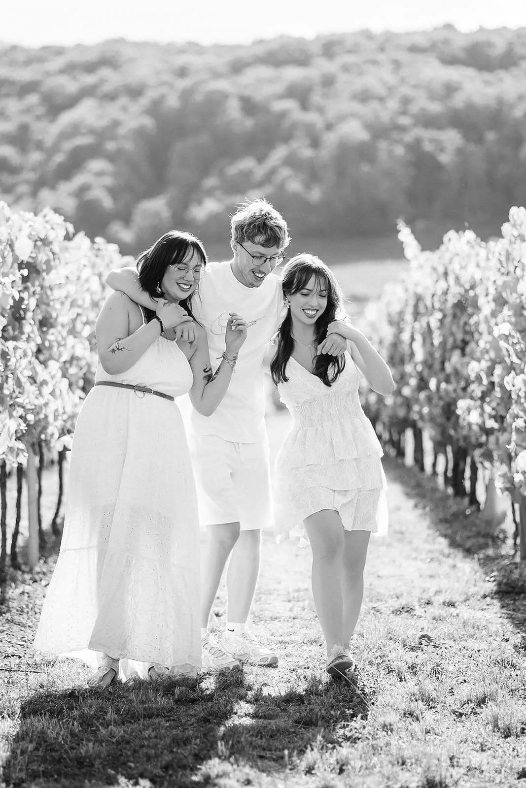 Une fratrie de trois jeunes adultes riant ensemble en marchant dans une allée de vignes à Boersch, photo noir et blanc