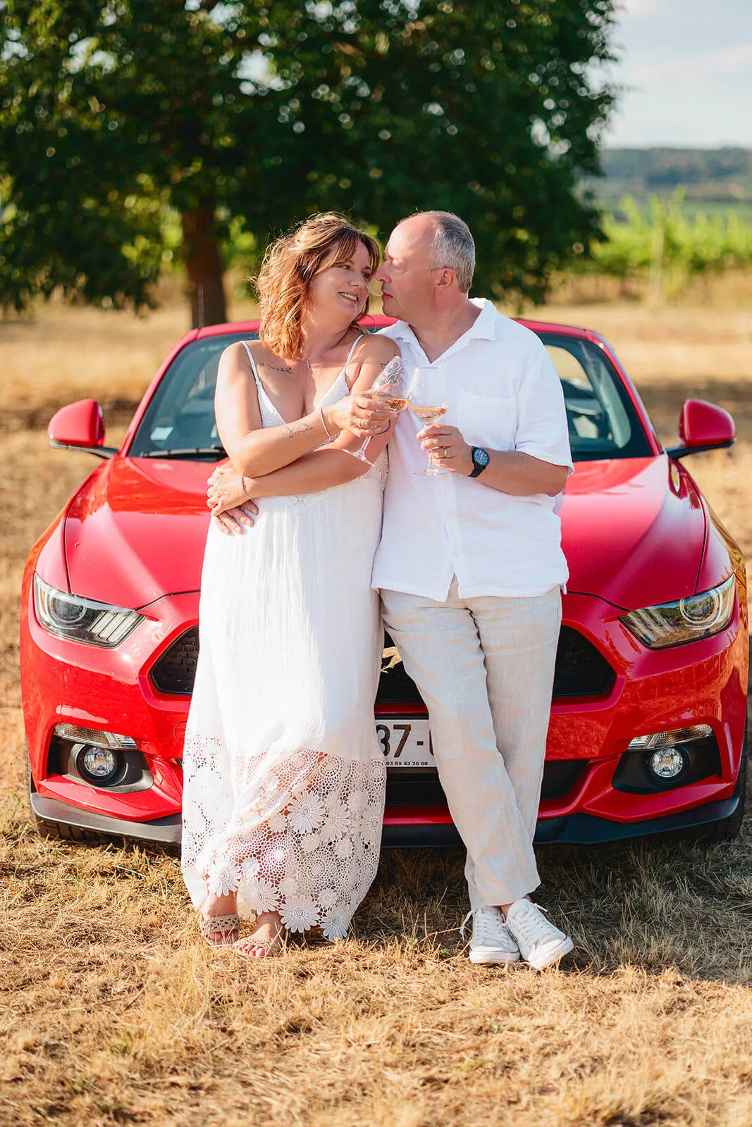 Couple trinquant avec un verre de vin devant leur voiture de sport rouge à Boersch
